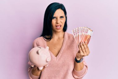 Norveç kronu banknotları ve domuz kumbarası suratlı, kuşkucu ve alaycı bakışlı, ağzı açık bir şekilde şaşırmış genç beyaz bir kadın. 