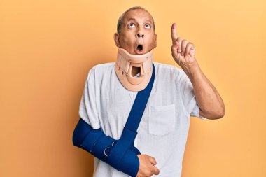 Servikal tasma ve kol takan yakışıklı, olgun bir adam. Şaşırmış ve şaşırmış bir şekilde yukarı bakıyor, parmaklarıyla işaret ediyor ve kollarını kaldırıyor.. 