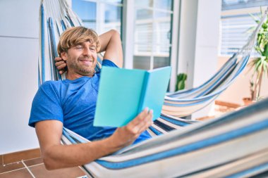 Orta yaşlı yakışıklı bir adam evinin terasında bir hamakta uzanıp kitap okuyor.