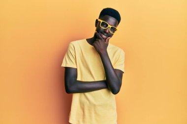 Sıradan kıyafetler ve gözlük takan genç Afro-Amerikalı adam kameraya kendinden emin bir şekilde bakıyor kollarını kavuşturmuş ve çenesini kaldırmış gülümsüyor. Olumlu düşünmek. 