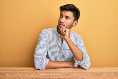 Genç ve yakışıklı bir adam, gündelik giysiler içinde masada oturmuş şüphe üzerine kafa yoruyor, parmakları çenesinde ve yukarı bakıyor. 
