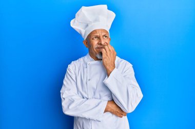 Profesyonel aşçı üniforması giyen ve şapkası olan orta yaşlı, gri saçlı bir adam gergin ve elleri ağızdan tırnak yerken gergin görünüyor. anksiyete. 