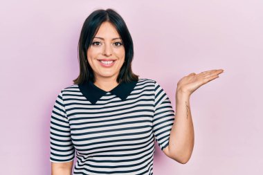 Gündelik kıyafetler giyen genç İspanyol kadın neşeli bir şekilde gülümsüyor ve avuç içiyle kameraya bakıyor.. 