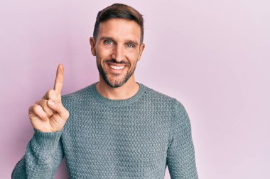 Kış süveteri giyen, sakallı yakışıklı bir adam bir fikir veya soruyla gülümseyen, mutlu bir yüzle işaret eden, bir numara 