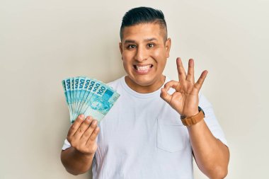 Elinde 100 Brezilyalı banknot tutan genç latin adam parmaklarıyla imza atıyor, gülümsüyor ve mükemmel bir sembol oluşturuyor. 