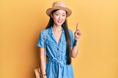 Genç Çinli kadın yaz şapkası takıyor ve bir numarayı gösterip işaret ederken kendinden emin ve mutlu gülümsüyor.. 