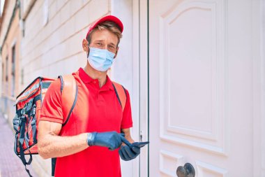 Beyaz postacı kırmızı üniforma ve teslimat çantası giyiyor ve akıllı telefon kullanarak koronavirüs güvenlik maskesi takıyor.