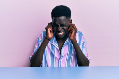 Genç Afro-Amerikan adam, gündelik giysiler içinde masaya oturmuş yüksek sesli müzik sesi için kulaklarını parmaklarıyla kapatıyor. sağır kavramı. 