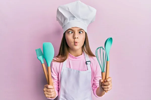 Beautiful brunette little girl wearing professional cook apron holding cooking tools making fish face with mouth and squinting eyes, crazy and comical.