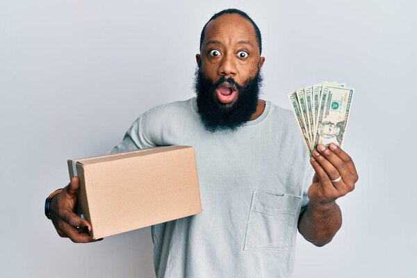 Молодой африканский американец с коробкой доставки и банкнотами в долларах США испугался и шокирован удивлением и удивлением выражения лица, страха и возбужденного лица. 