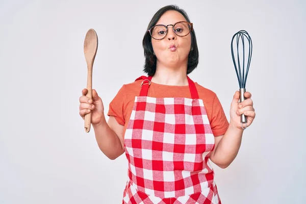 Brunette woman with down syndrome cooking using baker whisk and spoon making fish face with mouth and squinting eyes, crazy and comical.