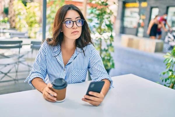 Young hispanic woman with serious expression using smartphone at coffee shop terrace.