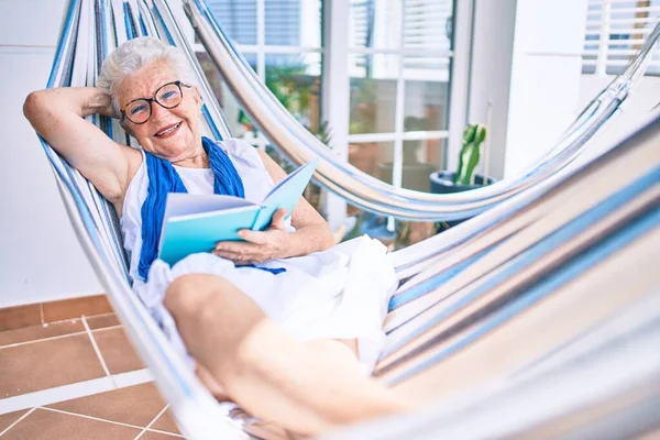 Elder senior woman with grey hair smiling happy relaxing on a hammock reading a book at home