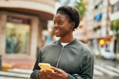Genç Afrikalı Amerikalı kız şehirde akıllı telefon kullanarak mutlu bir şekilde gülümsüyor.
