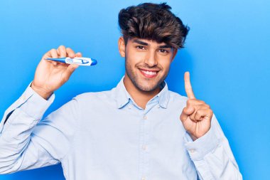 Elinde termometreyle gülümseyen genç İspanyol bir adam bir fikir ya da soruyla mutlu bir yüzle işaret ediyor. 