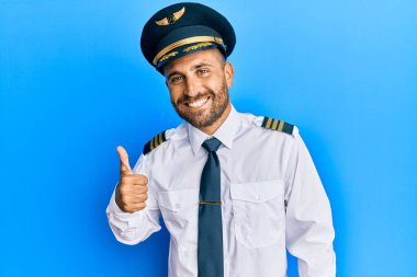 Uçak pilotu üniforması giyen sakallı yakışıklı el hareketleriyle mutlu bir jest yapıyor. Kameraya bakıp başarıyı gösteren ifade. 