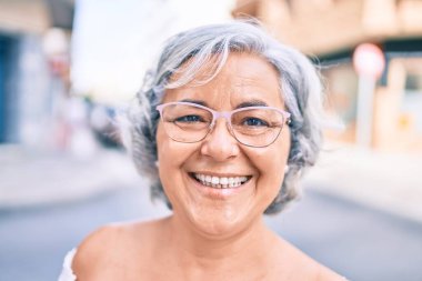 Beyaz saçlı orta yaşlı bir kadın mutlu bir şekilde dışarıda gülümsüyor.