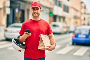 Beyaz atlı genç teslimatçı akıllı telefon kullanıyor ve şehirde kağıt torba taşıyor..