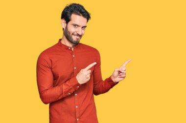 Gündelik kıyafetler giyen genç İspanyol adam gülümseyip kameraya bakıyor ve iki eliyle ve parmaklarıyla kenarı işaret ediyor.. 