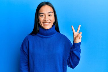 Genç Asyalı kadın sıradan bir süveter giyer ve iki numaralı parmaklarıyla işaret ederken kendinden emin ve mutlu bir şekilde gülümser.. 