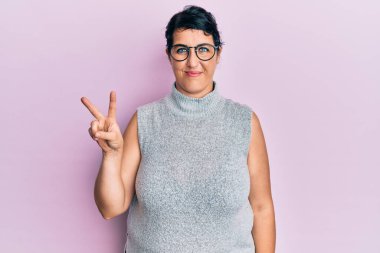 Artı, sıradan kıyafetler giyen ve gözlüklü esmer bir kadın kameraya gülümseyen mutlu bir yüzle zafer işareti yapıyor. İki numara.. 