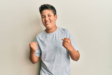 Gündelik gri tişört giyen genç İspanyol çocuk mutlu ve heyecanlı bir şekilde kolları havada, gülümseyen ve başarı için bağıran bir jest yapıyor. kutlama konsepti. 