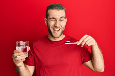 Diş macunuyla diş fırçası tutan genç beyaz bir adam kameraya seksi bir ifadeyle bakıyor. Neşeli ve mutlu bir yüzü var.. 