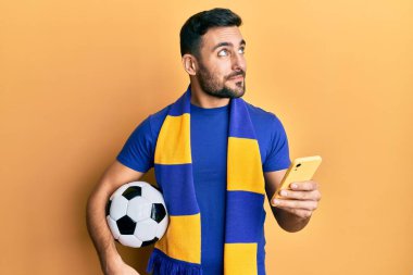 Genç İspanyol futbolcu holigan futbol topu tutuyor. Akıllı telefon kullanıyor. Gülümsüyor ve başka tarafa bakıyor.. 