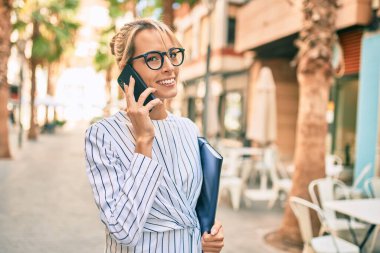 Sarışın bir iş kadınının elinde klasör var ve şehirdeki akıllı telefondan konuşuyor..