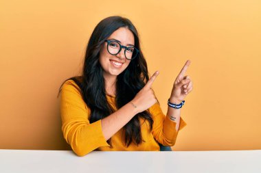 Güzel, esmer, gözlüklü, sıradan elbiseli bir kadın masaya oturmuş gülümsüyor ve kameraya bakıyor. İki eliyle ve parmaklarıyla işaret ediyor.. 