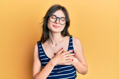 Gündelik kıyafetler ve gözlüklerle gülümseyen, elleri göğsünde, gözleri kapalı, yüzünde minnet dolu bir jestle genç, beyaz bir kız. sağlık kavramı. 