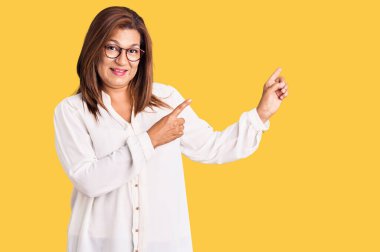 Orta yaşlı Latin kadın, günlük kıyafetler ve gözlük takıyor gülümsüyor ve kameraya bakıyor iki eliyle ve parmaklarıyla kenarı işaret ediyor.. 