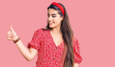 Yazlık elbise giyen esmer genç kız gururlu görünüyor, gülümsüyor yan tarafa baş parmak hareketi yapıyor. 