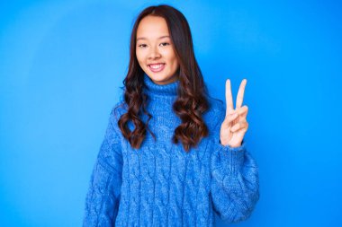 Genç, güzel, Çinli bir kız, sıradan bir kazak giyiyor. İki numaralı parmaklarıyla işaret ediyor. Kendinden emin ve mutlu bir şekilde gülümsüyor.. 