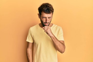 Sarı arka planda sarı tişört giyen sakallı yakışıklı adam kendini iyi hissetmiyor ve soğuk algınlığı veya bronşitin belirtisi olarak öksürüyor. sağlık hizmeti kavramı. 