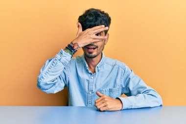 Gündelik giysiler giymiş genç İspanyol bir adam masaya oturmuş şok içinde yüzünü ve gözlerini kapayarak utanmış bir ifadeyle parmaklarının arasından bakıyor.. 