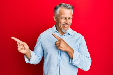 Orta yaşlı, gri saçlı, sıradan kıyafetler giyen, gülümseyen ve kameraya bakan, iki elini ve parmağını yana doğru gösteren bir adam.. 