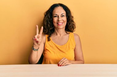 Masada oturan orta yaşlı, güzel bir kadın iki numaralı parmaklarını gösterip gülümserken kendinden emin ve mutlu görünüyor.. 