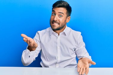 İş kıyafetleri giymiş yakışıklı İspanyol adam masaya oturmuş neşeli bir şekilde kollarını açıp gülümsüyor. Hoş geldin, olumlu ve kendinden emin selamlar. 