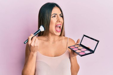 Makyaj fırçası tutan genç İspanyol kadın ve kızarık ve kızgın çığlıklar sinirli ve öfkeli, öfkeyle bağırıyor. öfke ve agresif konsept. 