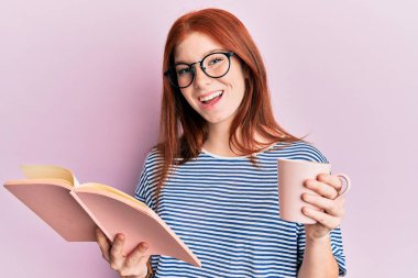 Genç kızıl saçlı kız kitap okuyor, bir fincan kahve içiyor ve kahkahalarla gülüyor.. 