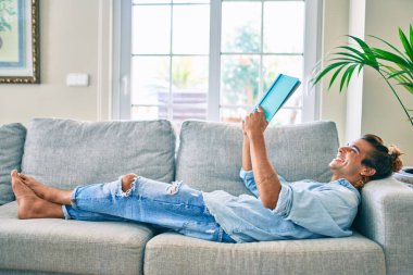 Genç İspanyol gülümseyen mutlu bir kitap okuyucu evdeki kanepede uzanıyor.