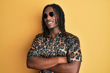 Saç örgülü, hayvan desenli tişörtlü ve güneş gözlüklü mutlu yüzlü, kollarını kavuşturup kameraya bakan Afro-Amerikan bir adam. pozitif kişi. 