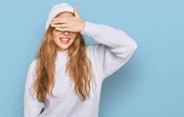 Genç beyaz kız yün kazak ve kış şapkası giyip gülümsüyor ve elleri yüzünde şaşkınlıktan gözleri kapayarak gülüyor. kör kavram. 