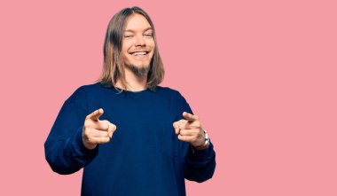 Uzun saçlı, kışlık süveter giyen yakışıklı beyaz adam kamerayı mutlu ve komik yüzüyle işaret ediyor. iyi enerji ve titreşimler. 