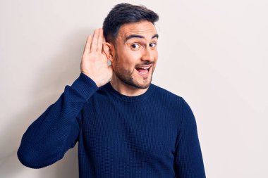 Sakallı, sıradan bir süveter giyen, beyaz arka planda dikilen, el pençe divan gülümseyen ve dedikoduları ve dedikoduları dinleyen genç yakışıklı bir adam. Sağırlık kavramı.