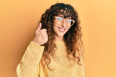 Günlük kıyafetler ve gözlük takan genç İspanyol kız mutlu ve pozitif gülümsüyor, başparmağıyla mükemmel ve onay işareti yapıyor. 