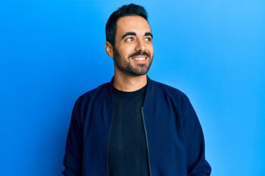 Latin kökenli genç bir adam günlük giysiler giyiyor, rahat bir görünüm takınıyor, doğal bir yüz ifadesi ve kendinden emin bir gülümsemeyle poz veriyor.. 