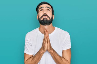 Latin asıllı genç bir adam, günlük beyaz t-shirt giyip elleri ile yalvarıyor ve dua ediyor. Yüzünde umut ifadesi var. Çok duygusal ve endişeli. Yalvarıyor. 