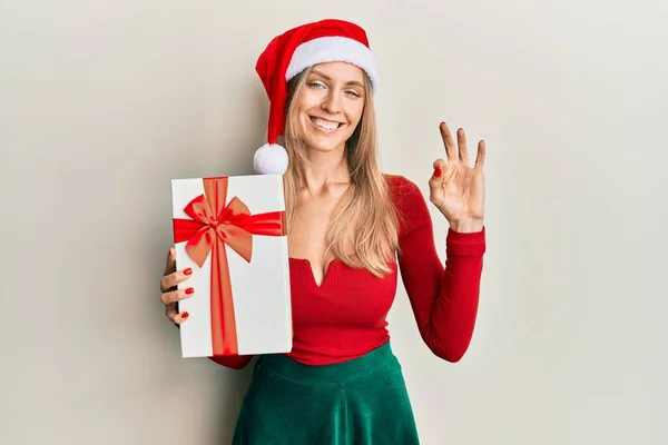 Beautiful Caucasian Woman Wearing Christmas Hat Holding Gift Doing Sign — Stock Photo, Image